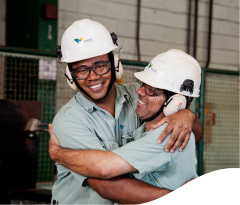 Foto da altura da barriga até a cabeça de dois empregados da Vale se abraçando e sorrindo. Os dois estão utilizando uniforme da Vale, camisa verde de botão, proteção nos ouvidos, óculos e capacete.