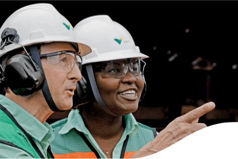 Shoulder to head shot of two Vale employees talking. The man is white and appears in the profile photo pointing his finger to a place. The woman is black, and she stands in front smiling. They are wearing helmets, goggles, and ear protection.