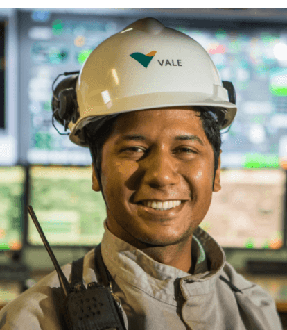 Vale employee smiling for a photo. He is wearing a protective helmet, and in the background you can see blurred computer screens.