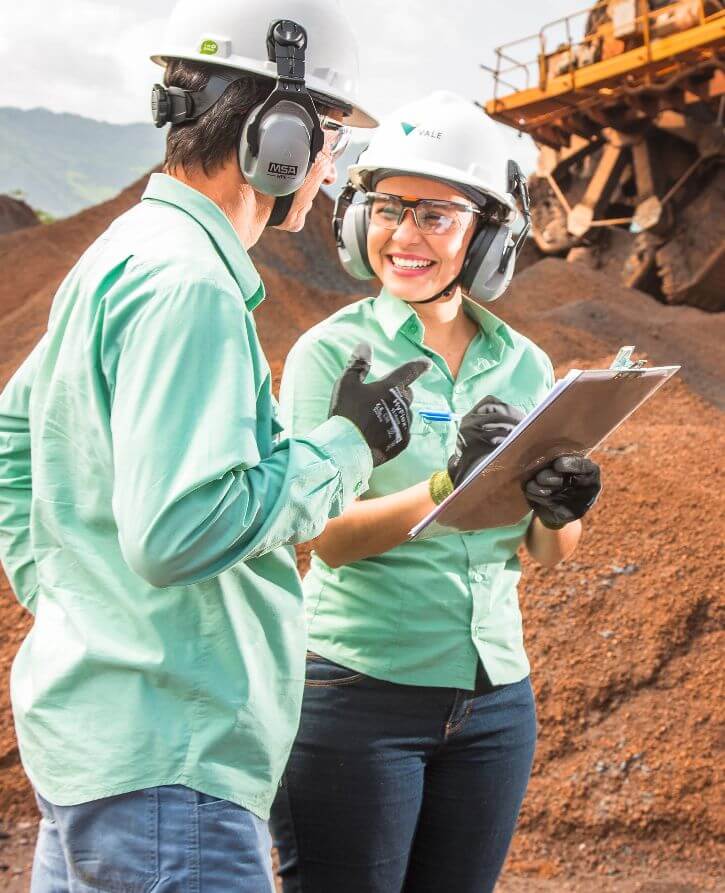 Two Vale employees – a man and a woman – standing side by side in an operations space with a large vehicle in the background. The two wear green shirts, gloves, goggles, earplugs, and white helmets. The woman holds a clipboard and smiles.