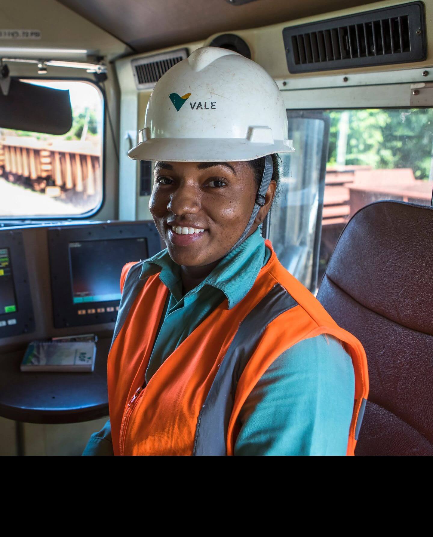 A black woman smiling inside a vehicle. She is wearing green Vale uniform, an orange vest and a white helmet with the company logo.