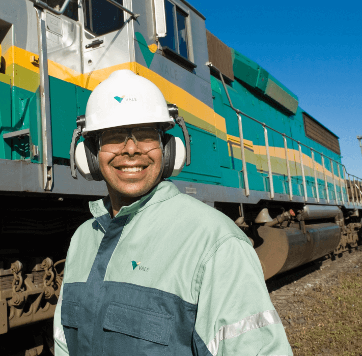 A man smiling next to a green, yellow and gray train. The man is wearing green uniform with Vale logo, goggles, ear muffs and white helmet with Vale logo.