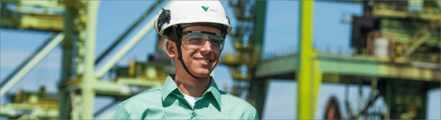 A man smiling in an operational area. He is wearing a light green shirt, goggles and a white helmet with Vale logo.