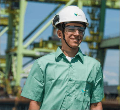 A man smiling in an operational area. He is wearing a light green shirt, goggles and a white helmet with Vale logo.