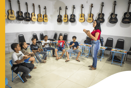 Music room with several guitars hanging on the wall. In the center, there are several children sitting in circles paying attention to a gesticulating woman.