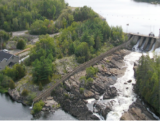 Vale-Per-28Photo of Wabageshik small hydropower plant with a concrete structure, rocks, vegetation, and moving water passing through the structures.