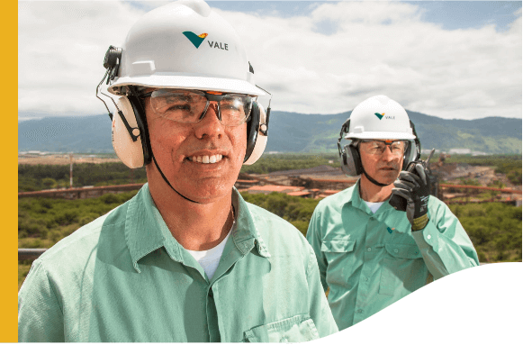 Photo of two men in a high part of an operation observing a point with vegetation, an operation and mountains in the background. One of the men is holding and talking on a radio with one hand. The other is smiling and watching. The two are wearing light green button-down shirts with Vale logo, gloves, ear protection, helmets and goggles.