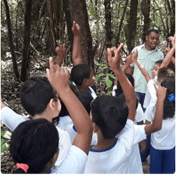 Several children, all wearing uniforms, are in a place with trees. In the background, it is possible to see Vale employee. The children are raising hands.