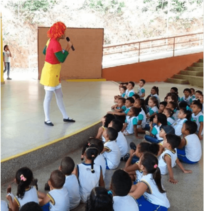 Em um palco, uma mulher está vestida de boneca de pano e fala em um microfone. Diversas crianças, de uniforme escolar, estão sentadas no chão prestando atenção na apresentação.