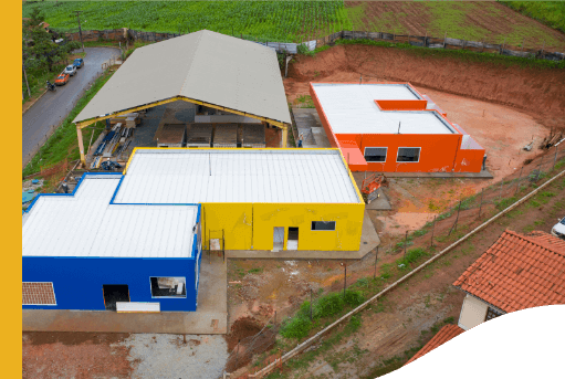 Aerial view of four colorful warehouses, the ground is dirt. In the background, it is possible to see a plantation.