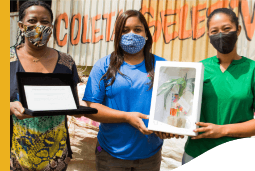 Three women, all wearing protective masks, pose for a photo. One of them is holding a certificate, and the other two, a box with a plant inside.