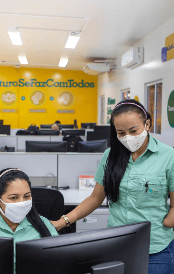 Two Vale female employees are side by side in an office. One is sitting in front of a computer and the other is standing. Both wear light green uniform shirts and white face masks