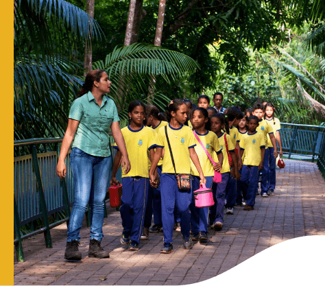 Vale female employee accompanies a big group of children, all dressed in school clothes, down a tree-lined path.