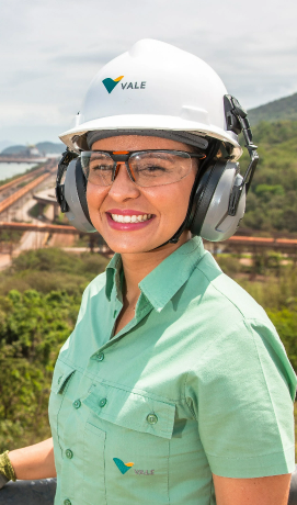 Foto da altura da cintura até a cabeça de uma mulher branca com uma operação ao fundo, usando uniforme da Vale: camisa botões verde, óculos, capacete e proteção nos ouvidos