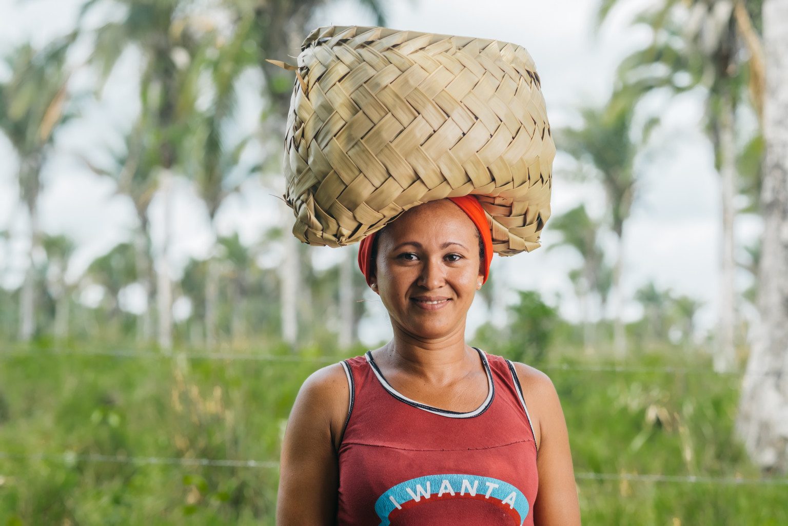  A woman balances a basket of straw on her head and smiles for a photo. In the background there are some trees