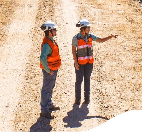 Photo seen from above, of the whole body of two Vale employees, talking in a place of dirt and sand, wearing uniform, gray pants, Green shirt, orange vest, helmet and glasses.
