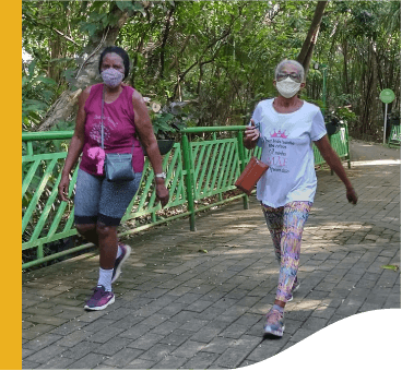 Duas senhoras caminham por um local arborizados. Ambas usam máscara de proteção.