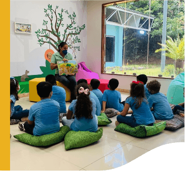 Enquanto uma empregada Vale mostra um livro, algumas crianças olham em direção a ela. As crianças estão sentadas em almofadas e usam uniformes azuis.