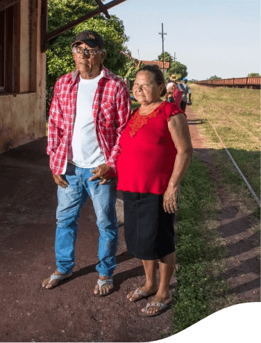 An old man and an old woman are standing in a grass place, where it is possible to see a train in the background He wears jeans, a white T-shirt, and a red plaid shirt, as well as glasses and a cap. She wears a black skirt and a red blouse. Both are wearing slippers.