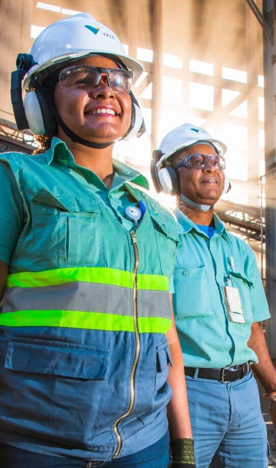 Two Vale employees – a man and a woman – standing next to each other in an operation area. The two are smiling and wear green clothing, goggles, ear muffs and white helmets with Vale logo.