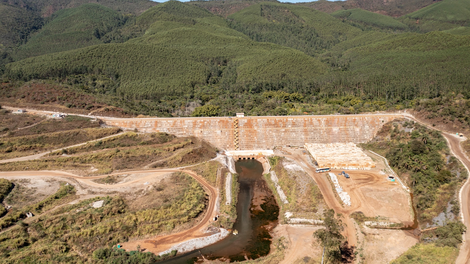 Construção de barragem cercada com área arborizada e com vegetação viva ao fundo.