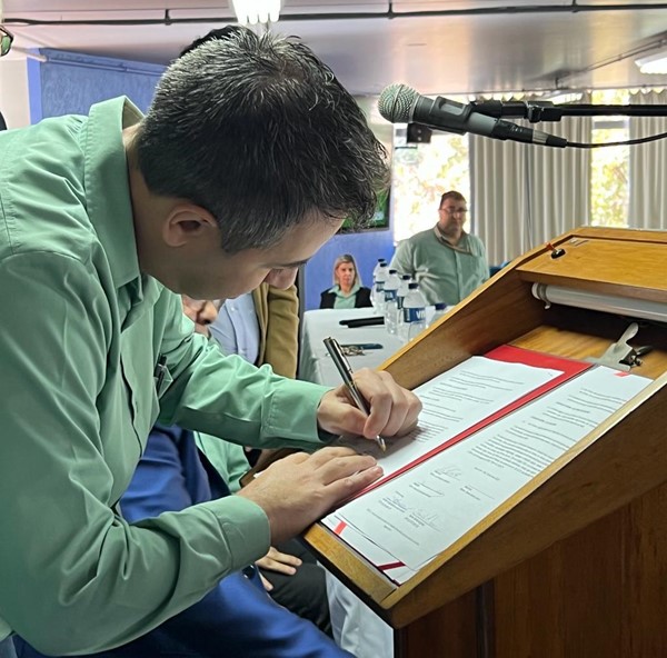 Homem usando blusa de manga longa segura caneta esferográfica com mão esquerda para assinar documento enquanto outras pessoas assistem.