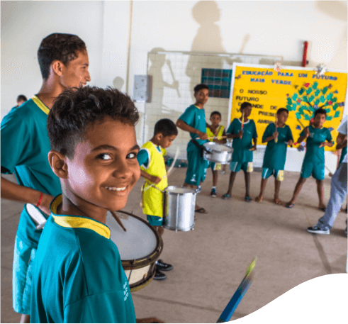 Várias crianças reunidas tocando instrumentos musicais em uma quadra. Todas elas usam roupas verdes e um menino olha para a câmera sorrindo.