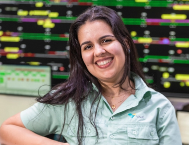 Photo of a woman in an office with multiple monitors in the background. She's leaning against a counter and is wearing a green button-down shirt with Vale's logo, long straight hair and is smiling.