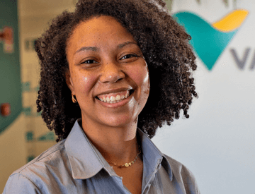 A black woman standing in an office space, wearing a gray shirt and smiling.