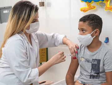 Foto de uma profissional da saúde segurando o braço de uma criança com uma das mãos e aplicando uma injeção com a outra. Ambas usam máscara facial.