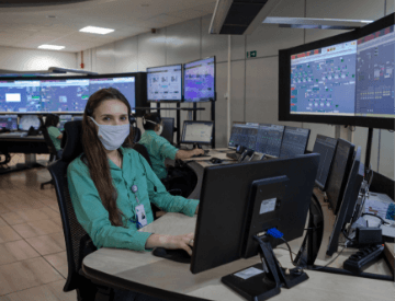 Photo of a woman sitting on a chair working on a computer in an office with several monitors and screens in the background. She has long dark hair and is wearing a uniform, a green button-up blouse, badge, and face mask.