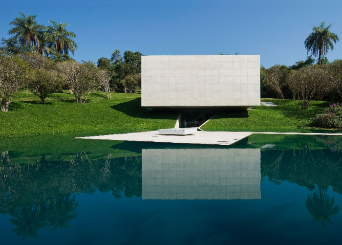 Vista externa da Galeria Adriana Varejão, Instituto Inhotim. Tacoa Arquitetos. Foto: Rossana Magri. O espaço é gramado e possui um lago que reflete a galeria. A estrutura da galeria é retangular e de concreto.